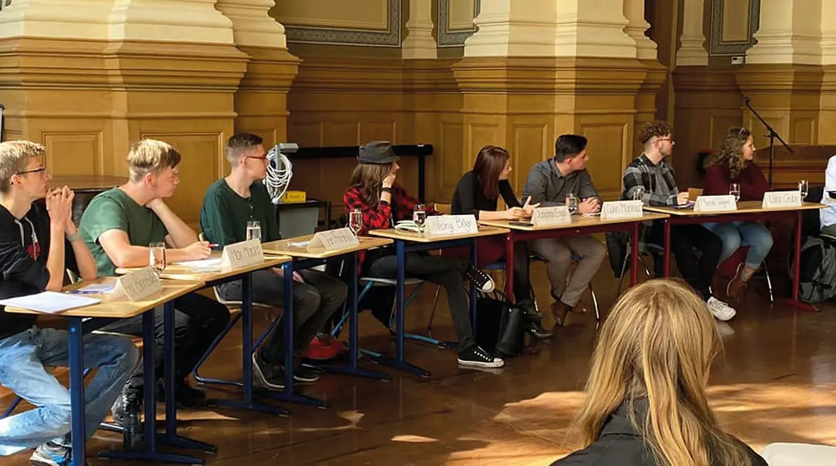 Mehrere Jugendliche sitzen an einem langen Tisch in einem Saal