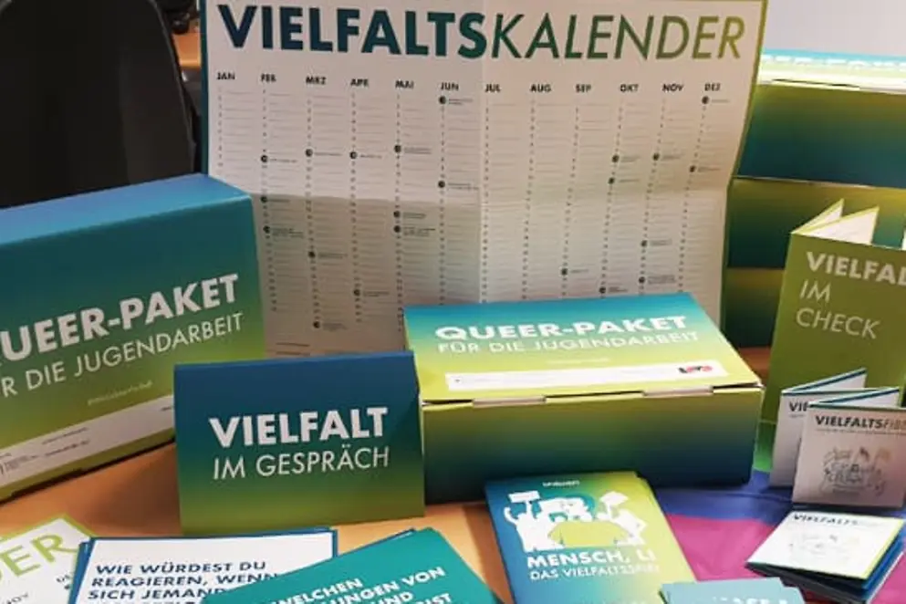 Auf einem Tisch liegen Broschüren, Kalender, Karten, Pappkisten und eine Regenbogenflagge