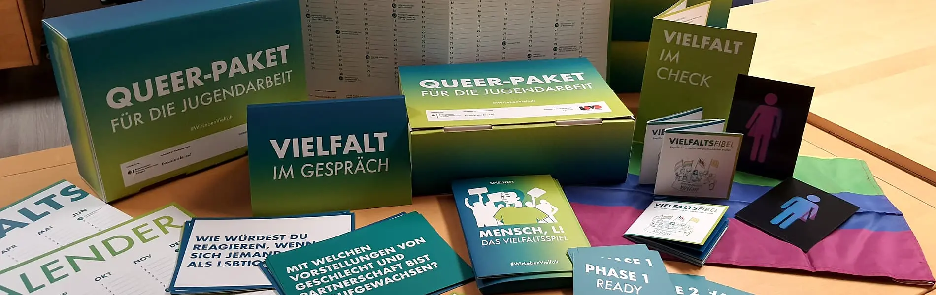 Auf einem Tisch liegen Broschüren, Kalender, Karten, Pappkisten und eine Regenbogenflagge