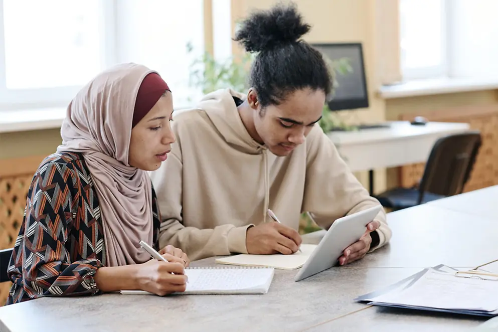 Zwei Personen an einem Tisch schauen in ein Tablet und schreiben auf Blöcke