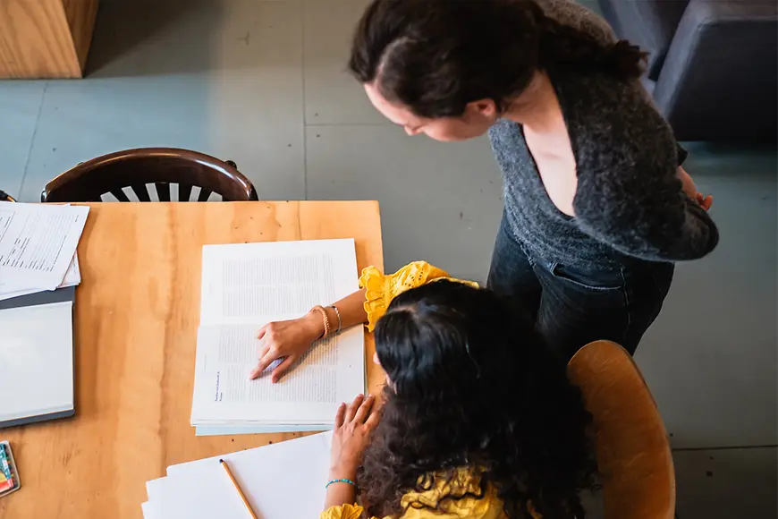 Eine Frau schaut einer anderen Frau über die Schulter, die an einem Tisch sitzt und liest.