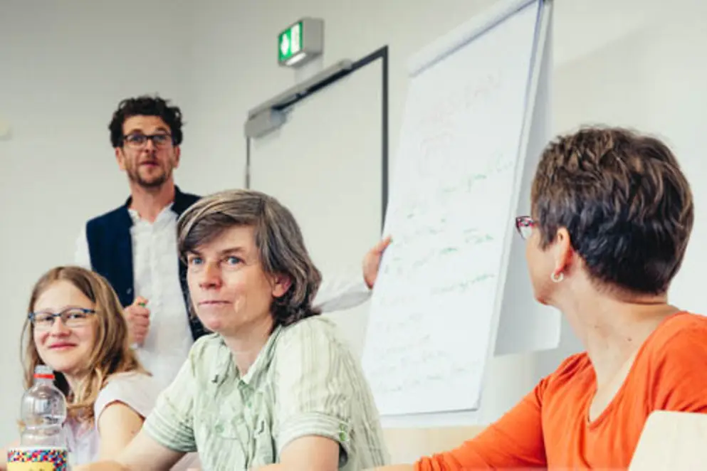 Drei Personen sitzen, eine Person steht an einem Flipchart