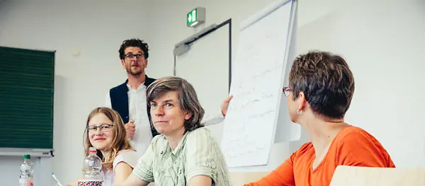 Drei Personen sitzen, eine Person steht an einem Flipchart