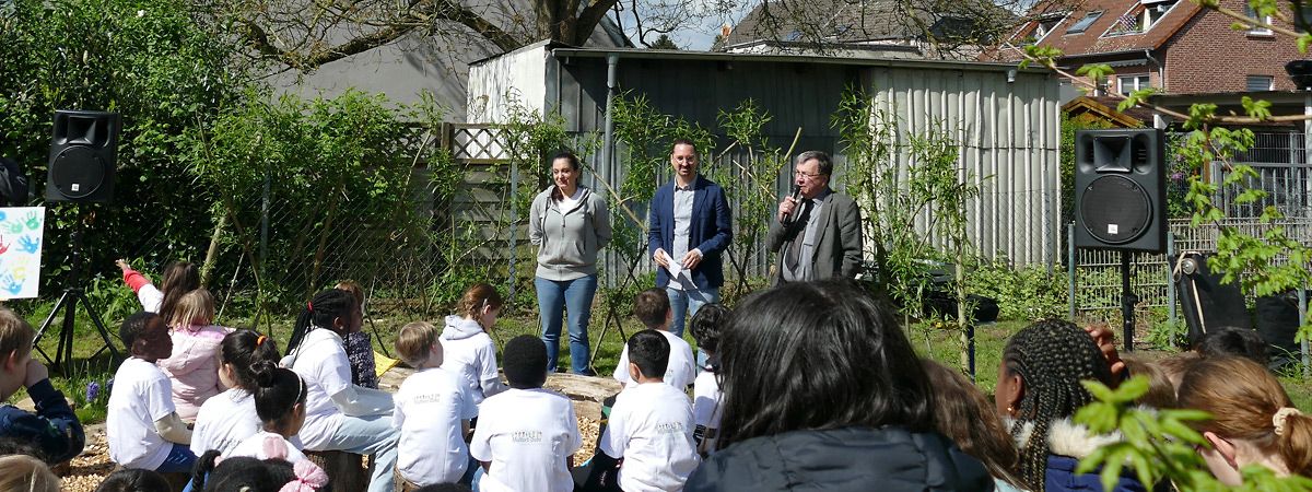 Grundschulgarten: Viele Kinder schauen auf drei, der Kamera zugewandeten, Menschen