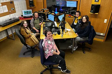 Fünf Personen sitzen in einem Aufnahmestudio und schauen den Betrachter an.