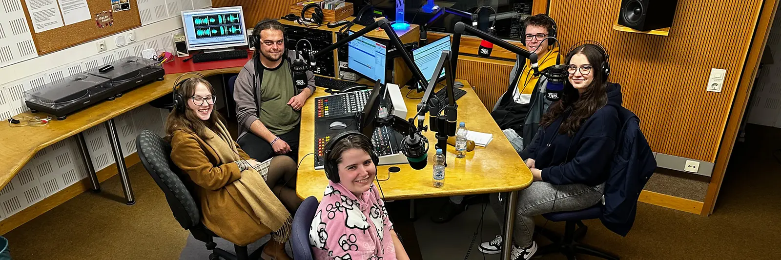 Fünf Personen sitzen in einem Aufnahmestudio und schauen den Betrachter an.